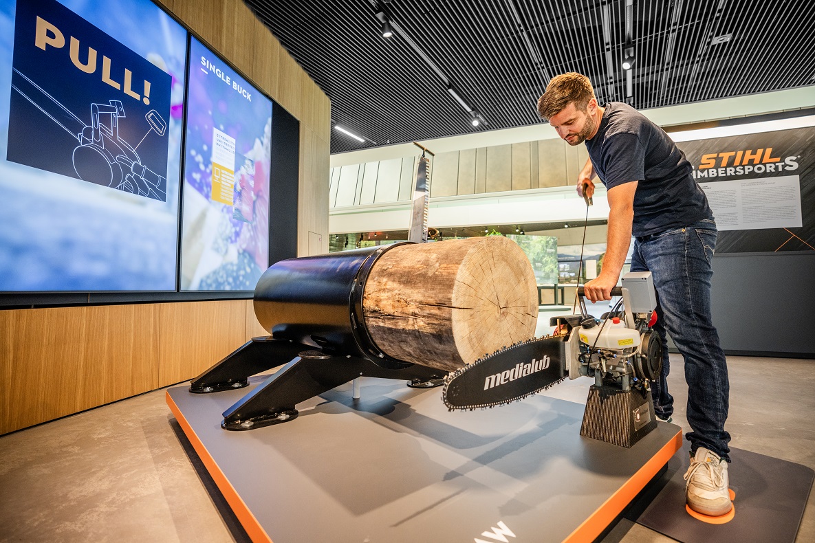 A man holds a chainsaw in his hand in front of a log in the STIHL Brand World.